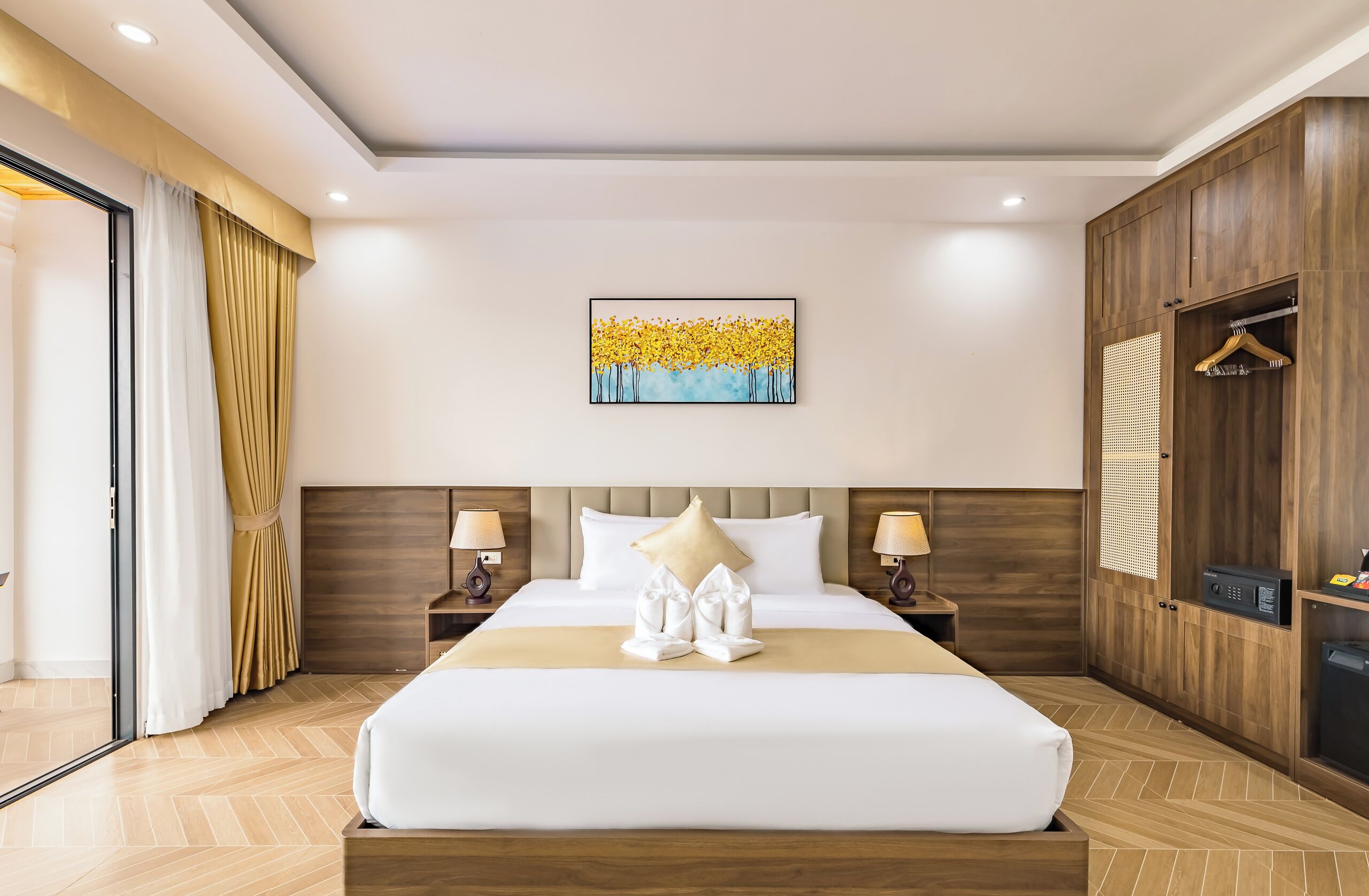 A hotel room with a large bed, wooden headboard, two bedside tables with lamps, a wall-mounted artwork, and a wardrobe with a hanging rack.