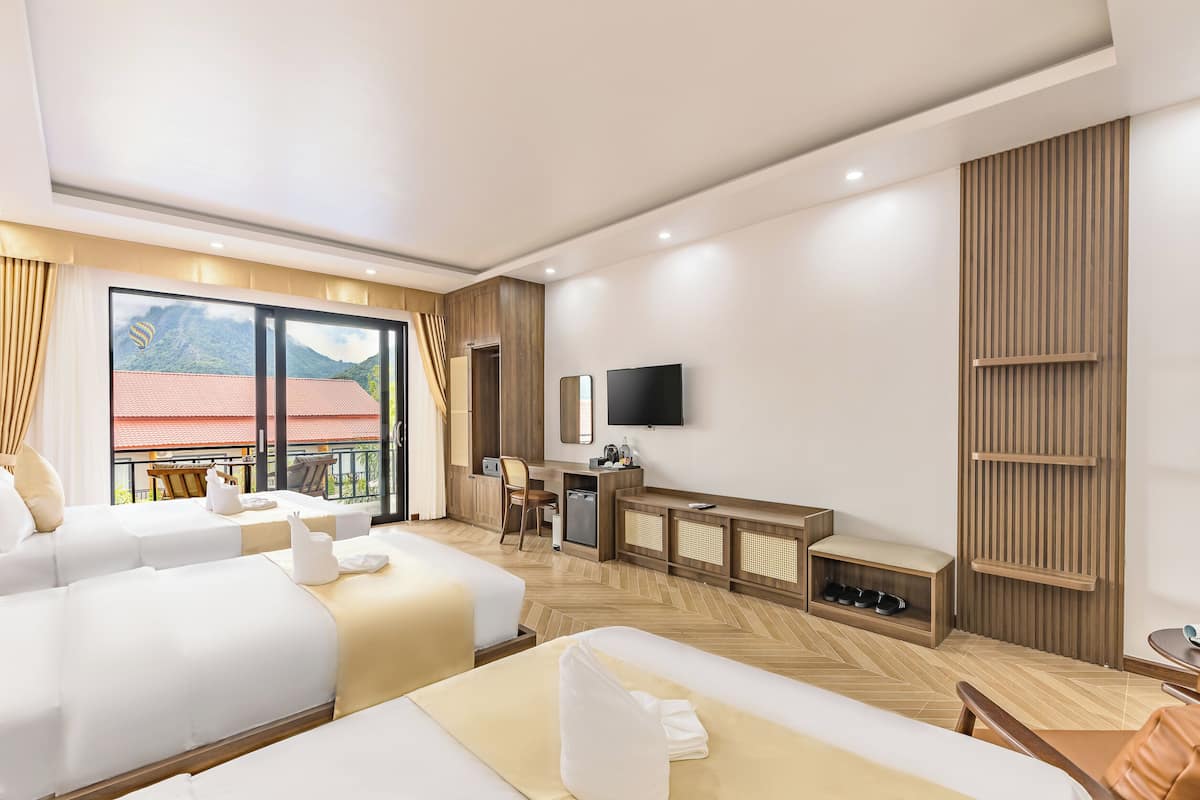A hotel room with two beds, a desk with a TV, a balcony with a mountain view, and a wooden floor.