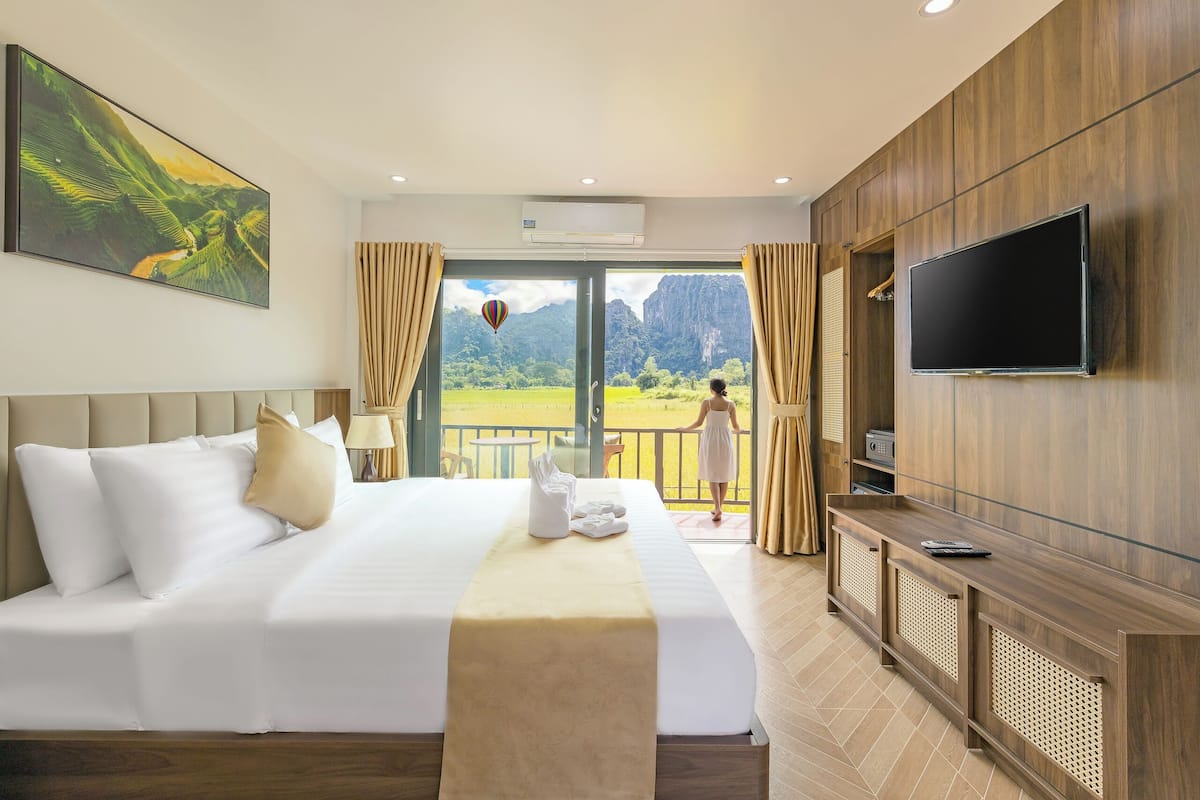 A hotel room with a bed, a TV, a balcony with a view of mountains, and a painting on the wall.