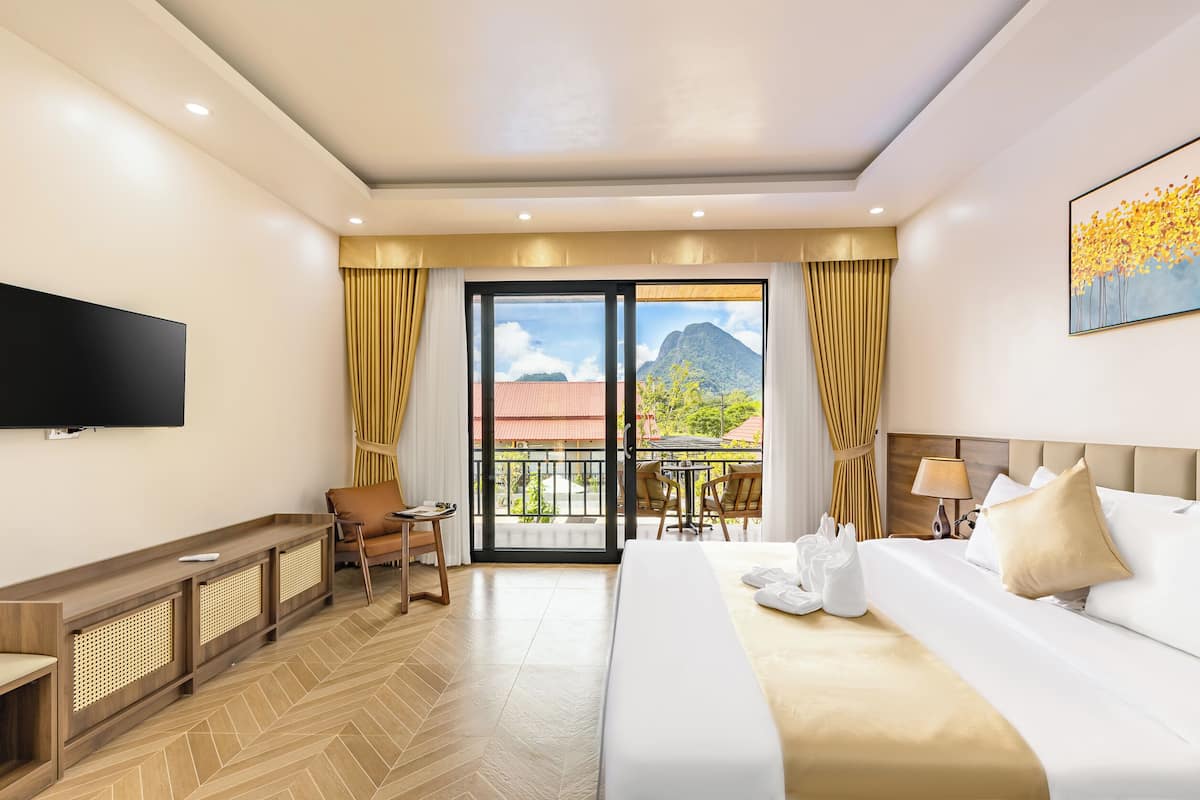 A hotel room with a bed, a TV, a balcony with a table and chairs, and a view of a mountain.