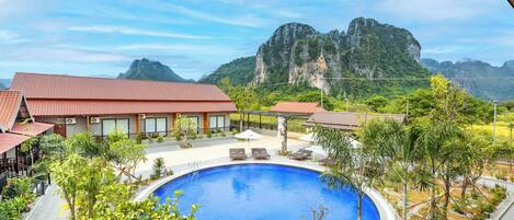 A hotel with a swimming pool surrounded by greenery and a view of a mountainous landscape.
