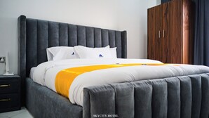 A modern bedroom with a large bed, grey upholstered headboard, white bedding, and a wooden wardrobe. - Skycity Hotel Gh (Amrahia)