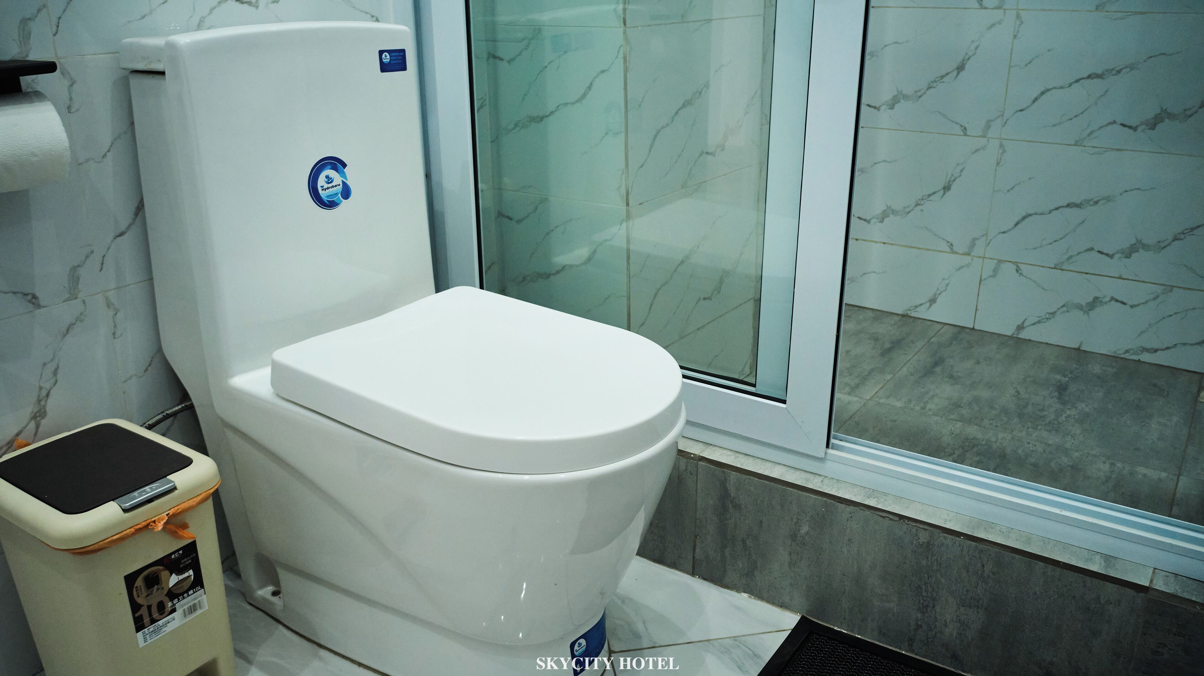 A white toilet with a blue sticker, a trash bin, and a shower area with marble tiles.