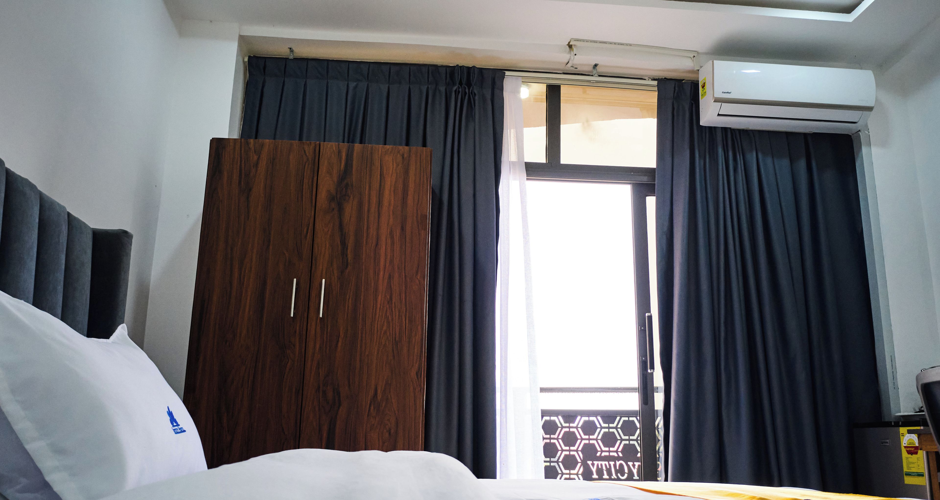 A hotel room with a bed, a wardrobe, a window with curtains, and an air conditioning unit.