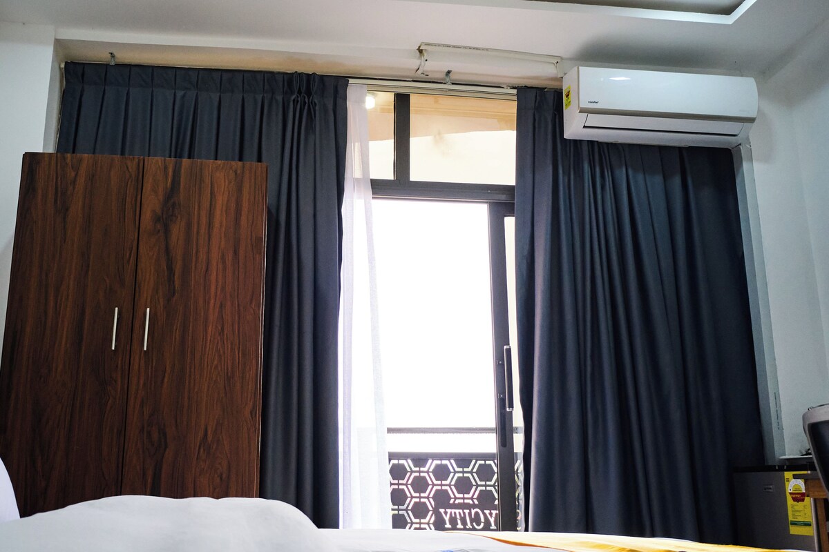 A hotel room with a bed, a wardrobe, a window with curtains, and an air conditioning unit.