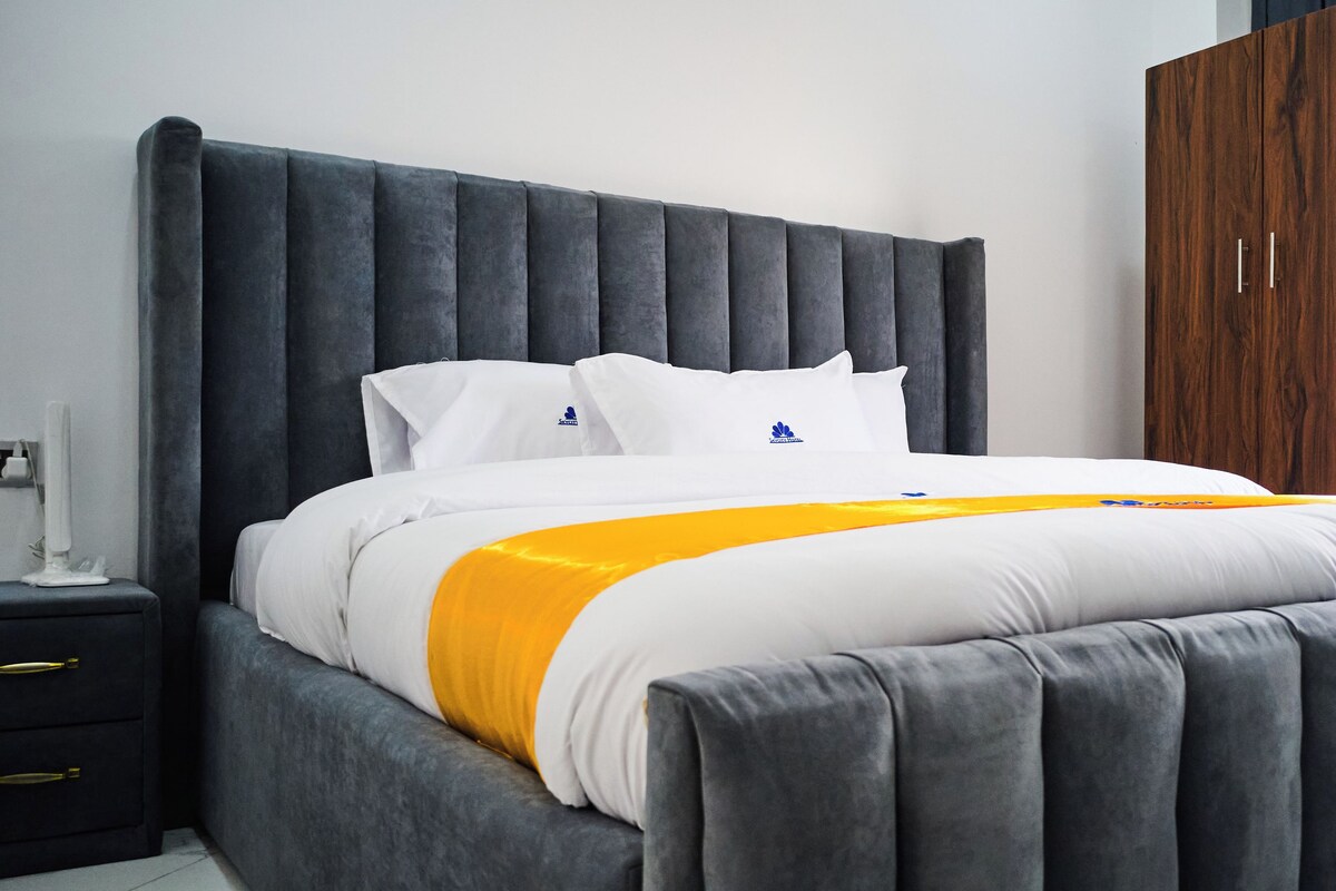 A modern bedroom with a large bed, grey upholstered headboard, white bedding, and a wooden wardrobe.