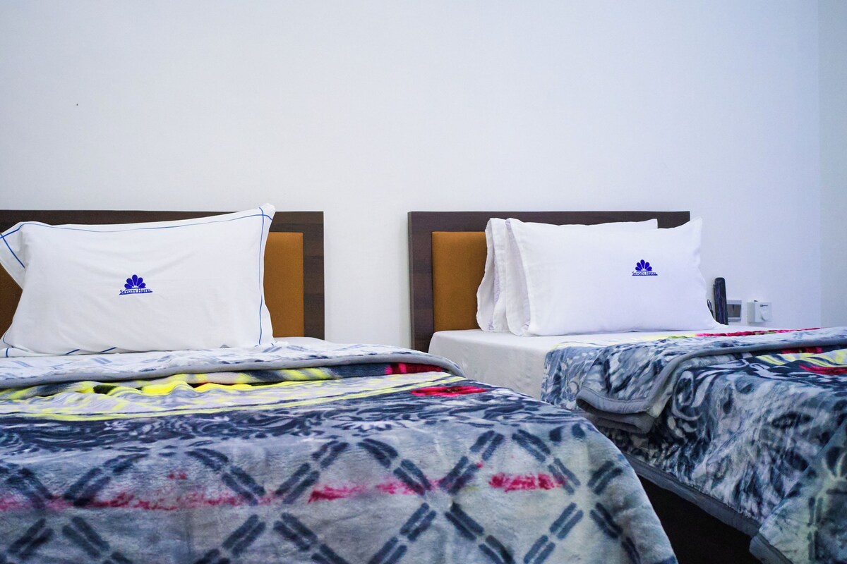 Two beds in a hotel room with headboards, white pillows with logos, and patterned bedspreads.