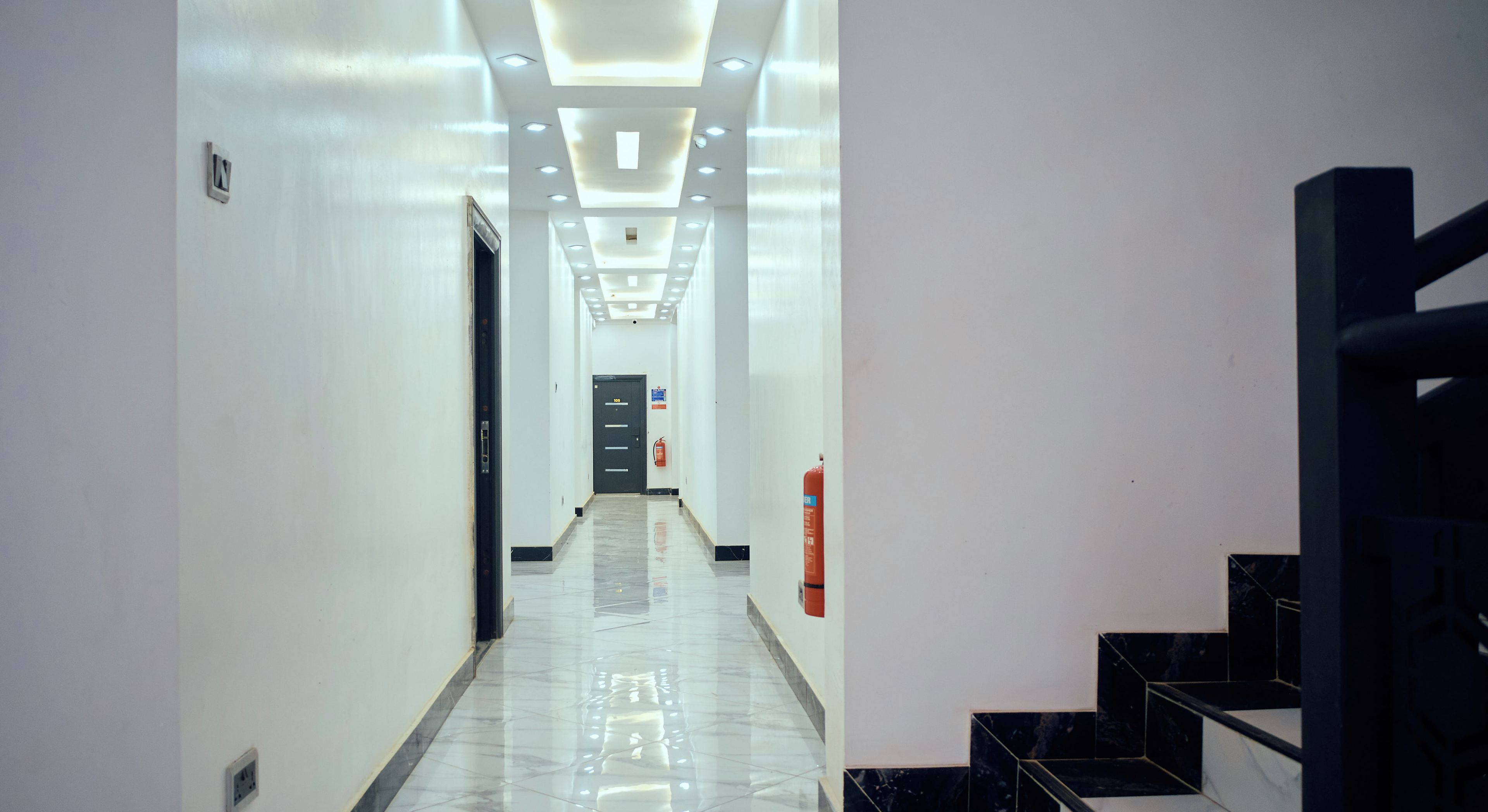 Un couloir d’hôtel au design moderne, avec un long couloir doté d’un sol en marbre, un escalier sur la droite et une porte au bout.
