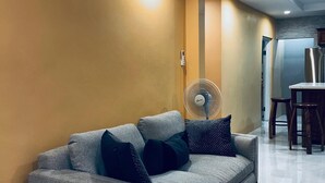 A modern living room with a grey sofa, a standing fan, and a dining area with wooden chairs and a table. - Jebucks Escape (Portmore)