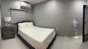 A bedroom with a bed, bedside table, lamp, air conditioning unit, and a standing fan. - Jebucks Escape (Portmore)