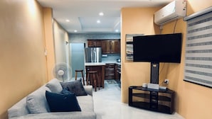 A modern living room with a grey sofa, a flat-screen TV mounted on a black stand, and a kitchen in the background. - Jebucks Escape (Portmore)