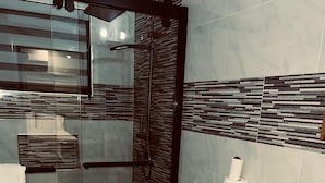 A modern bathroom with a glass shower enclosure, a toilet with a paper towel holder, and a tiled backsplash. - Jebucks Escape (Portmore)