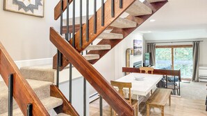 Uma escadaria moderna com degraus de madeira e corrimãos de metal, que leva a uma área de jantar com uma mesa e cadeiras brancas.