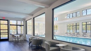 Uma área interna de piscina moderna, com amplas janelas, cadeiras de lounge cinzas e um espaço de estar junto à piscina.