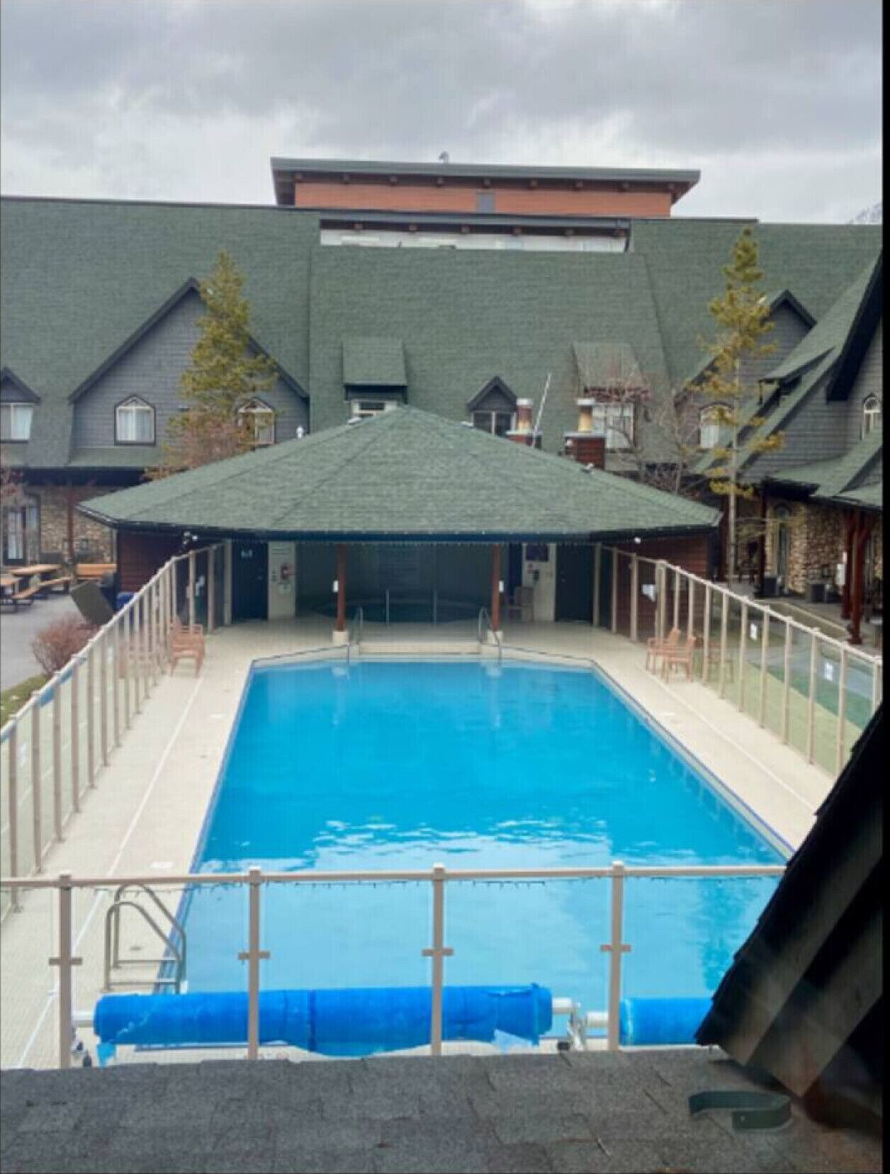 A rectangular swimming pool with a covered area and a roof, surrounded by a fence and a stone building in the background.