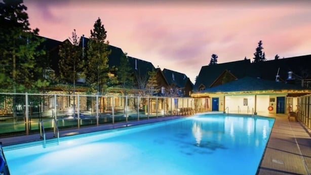Piscina com cobertura de vidro, cercada por edifícios e árvores, sob um céu crepuscular.