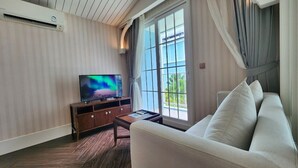 A living room with a sofa, a TV on a wooden cabinet, and a large window with curtains. - Grand Florida 1Bedroom CloseToBeach G410 (Sattahip)