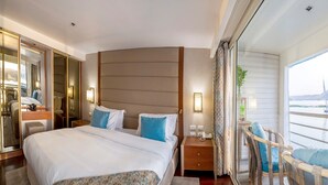 A hotel room with a large bed, a nightstand, a mirror, a window with a view of a body of water and a sailboat, and a chair by the window. - Sonesta Sun Goddess Nile cruise ship - 4 & 7 nights - From Luxor every Monday (Luxor)