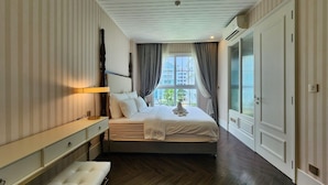 A bedroom with a large bed, a bedside table, a bench, a window with curtains, and a view of buildings outside.