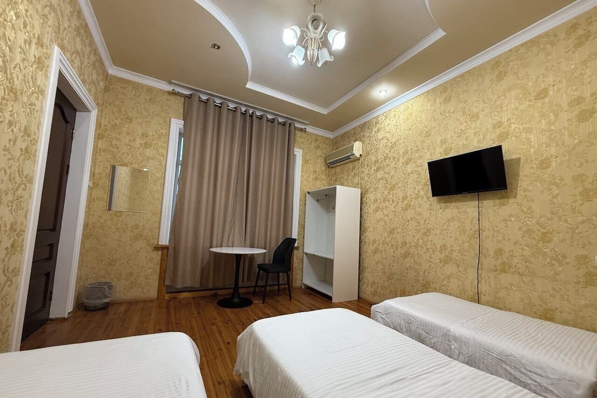 A hotel room with two single beds, a small table, a chair, a wardrobe, a television, and an air conditioning unit.