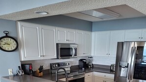 Cozinha moderna com armários brancos, eletrodomésticos de aço inoxidável, ilha de madeira com banquetas e um relógio na parede.
