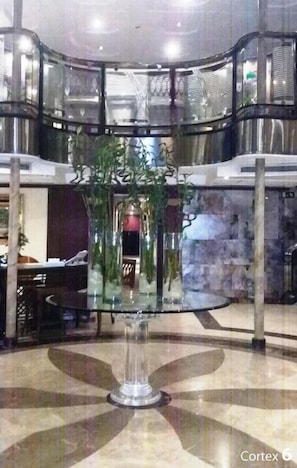 A glass-top table with a vase of green plants in a modern lobby with a circular floor pattern and a high ceiling.