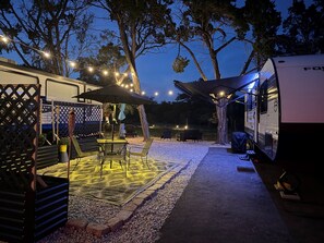 An RV campsite with a covered patio area, outdoor seating, and string lights. - Moonlite Wolf RV: King, Bunks, Fire Pit & Outdoor! (Spring Branch)