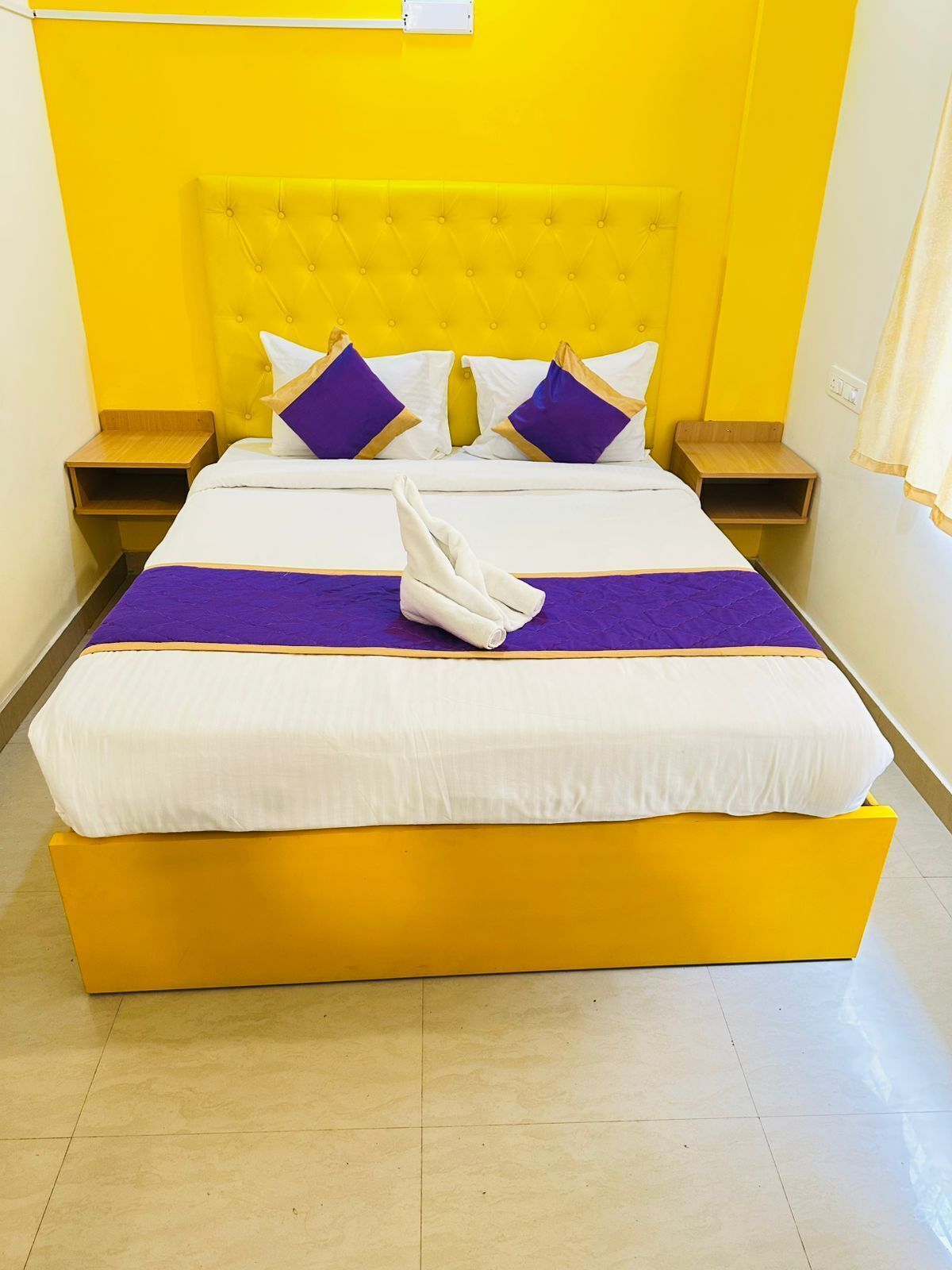 Una cama con cabecera amarilla, ropa de cama blanca y morada, y una toalla doblada en el pie de la cama.