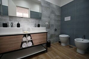 A modern bathroom with two sinks, a toilet, and a bidet. There are towels stored in a wooden cabinet. - Kore suites&apartments (Locri)