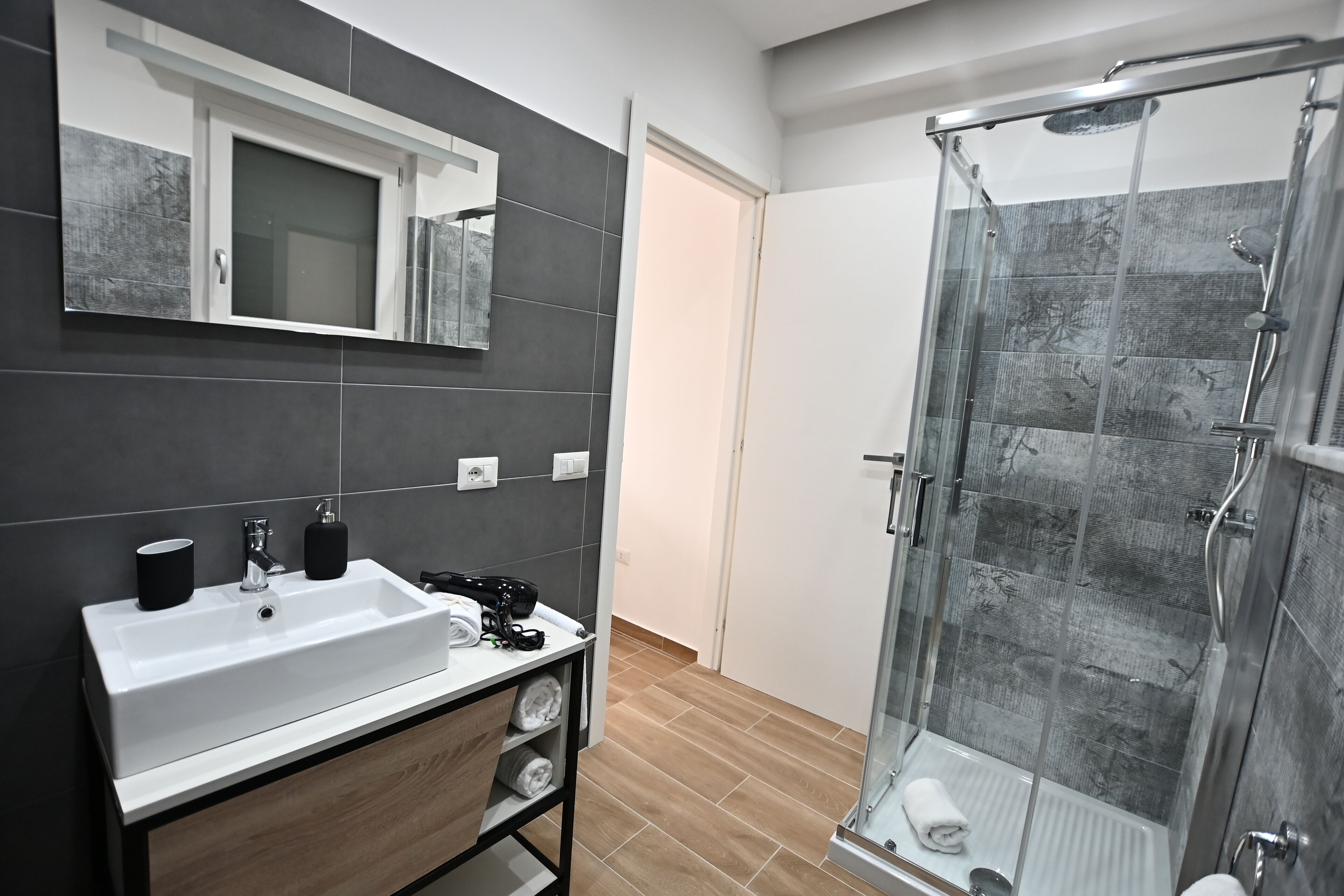 Une salle de bain moderne avec un lavabo blanc, des carreaux foncés, une douche en verre et un meuble-lavabo en bois.