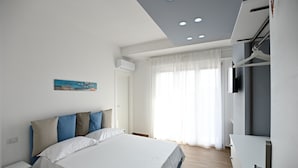 A modern hotel room with a large bed, a flat-screen TV, and a window with sheer curtains. - Kore suites&apartments (Locri)