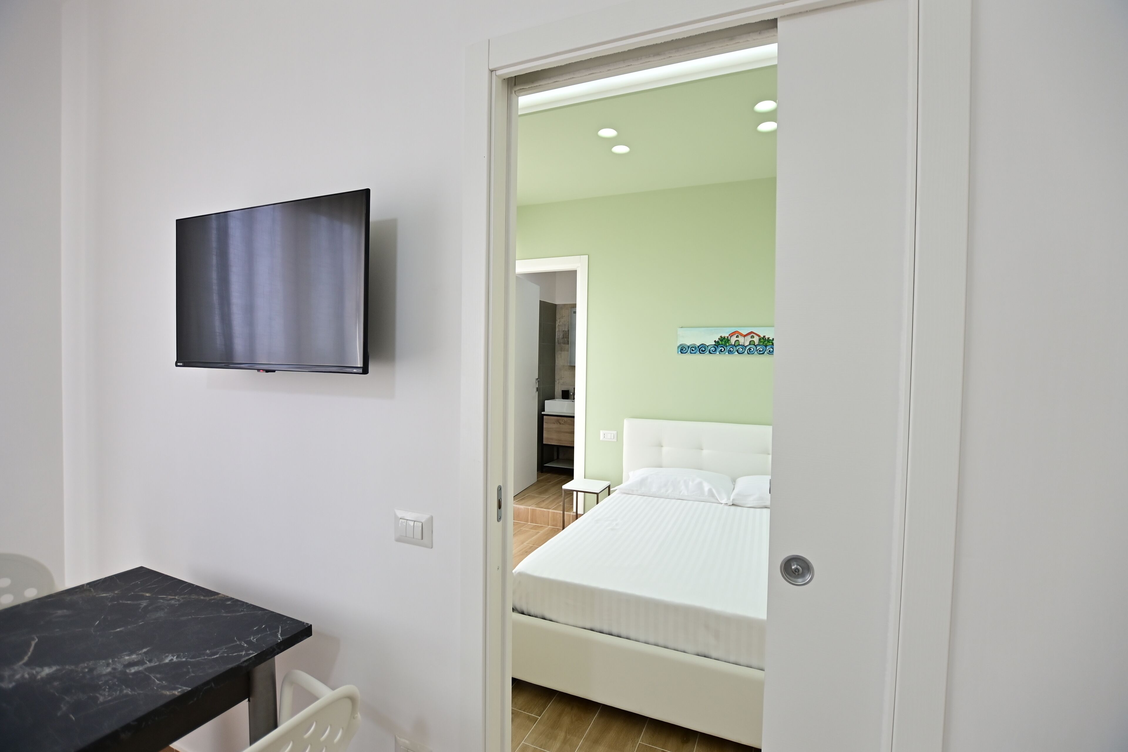 Una habitación de hotel con una cama, un televisor montado en la pared, una mesita y una puerta que da a otra habitación.