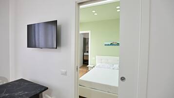 Una habitación de hotel con una cama, un televisor montado en la pared, una mesita y una puerta que da a otra habitación.