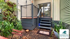 Una terraza de madera con barandilla metálica, un conjunto de escalones que conducen a una puerta y una planta en maceta junto a la puerta.