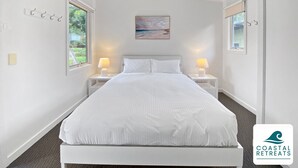 Un dormitorio blanco con una cama, dos mesitas de noche con lámparas, un cuadro en la pared y una ventana a la izquierda.