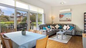 Amplia sala de estar con mesa de comedor, sofá gris con cojines de colores y un gran ventanal con vistas al exterior.