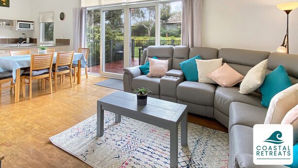 Una sala moderna con un sofá gris, una mesa de comedor de madera y sillas. Hay un ventanal con cortinas, un ventilador de techo y una lámpara.