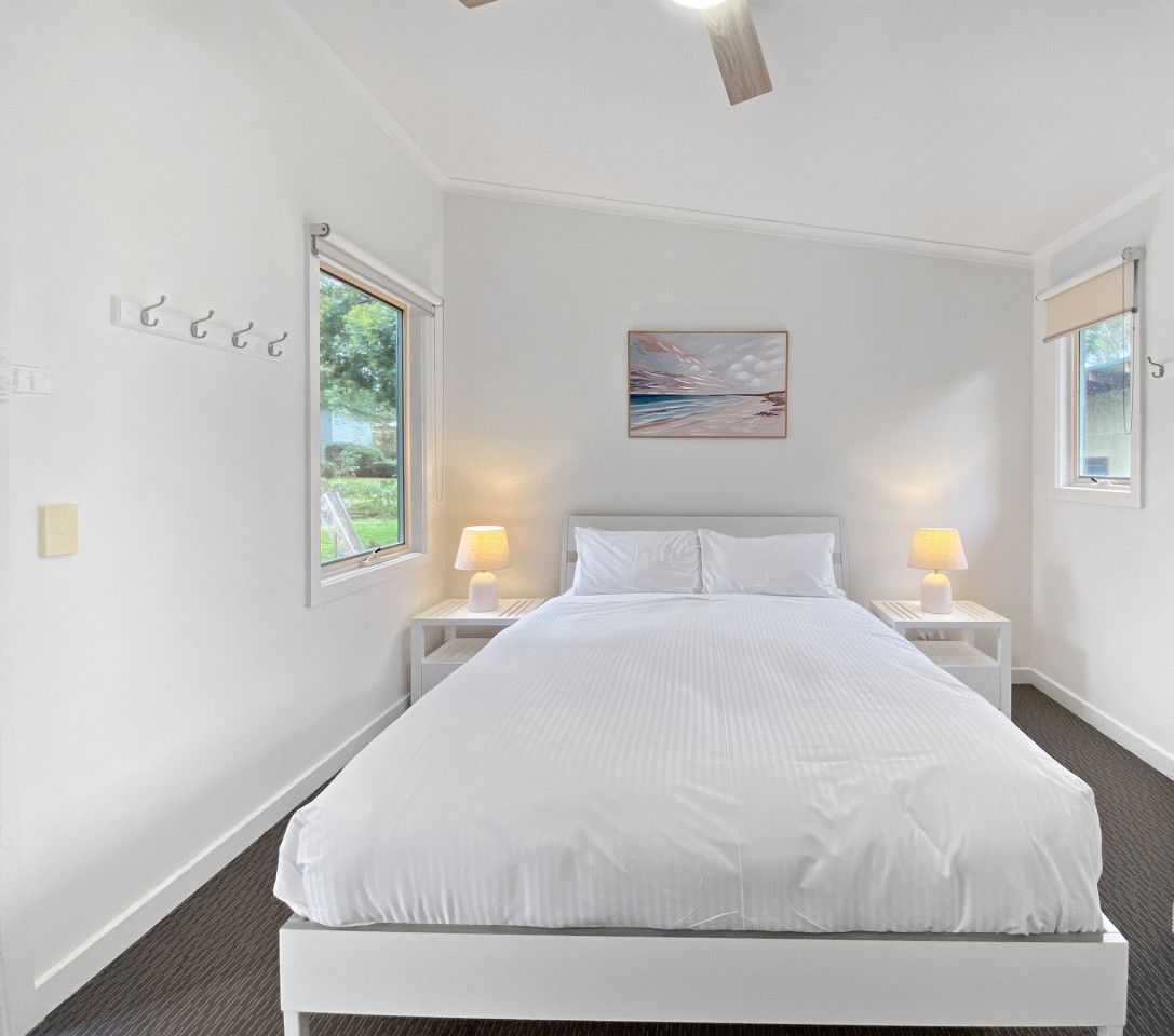 Une chambre à coucher blanche avec un lit, deux tables de chevet avec des lampes, un tableau au mur et une fenêtre sur le côté gauche.