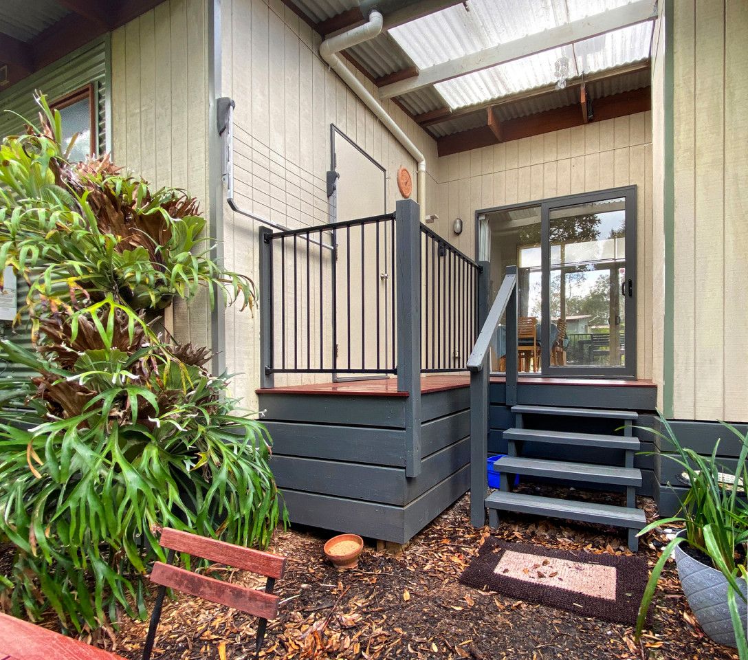 Une terrasse en bois avec une rambarde en métal, un escalier menant à une porte et un pot de fleurs placé à côté de la porte.