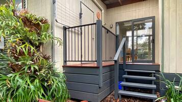 Une terrasse en bois avec une rambarde en métal, un escalier menant à une porte et un pot de fleurs placé à côté de la porte.