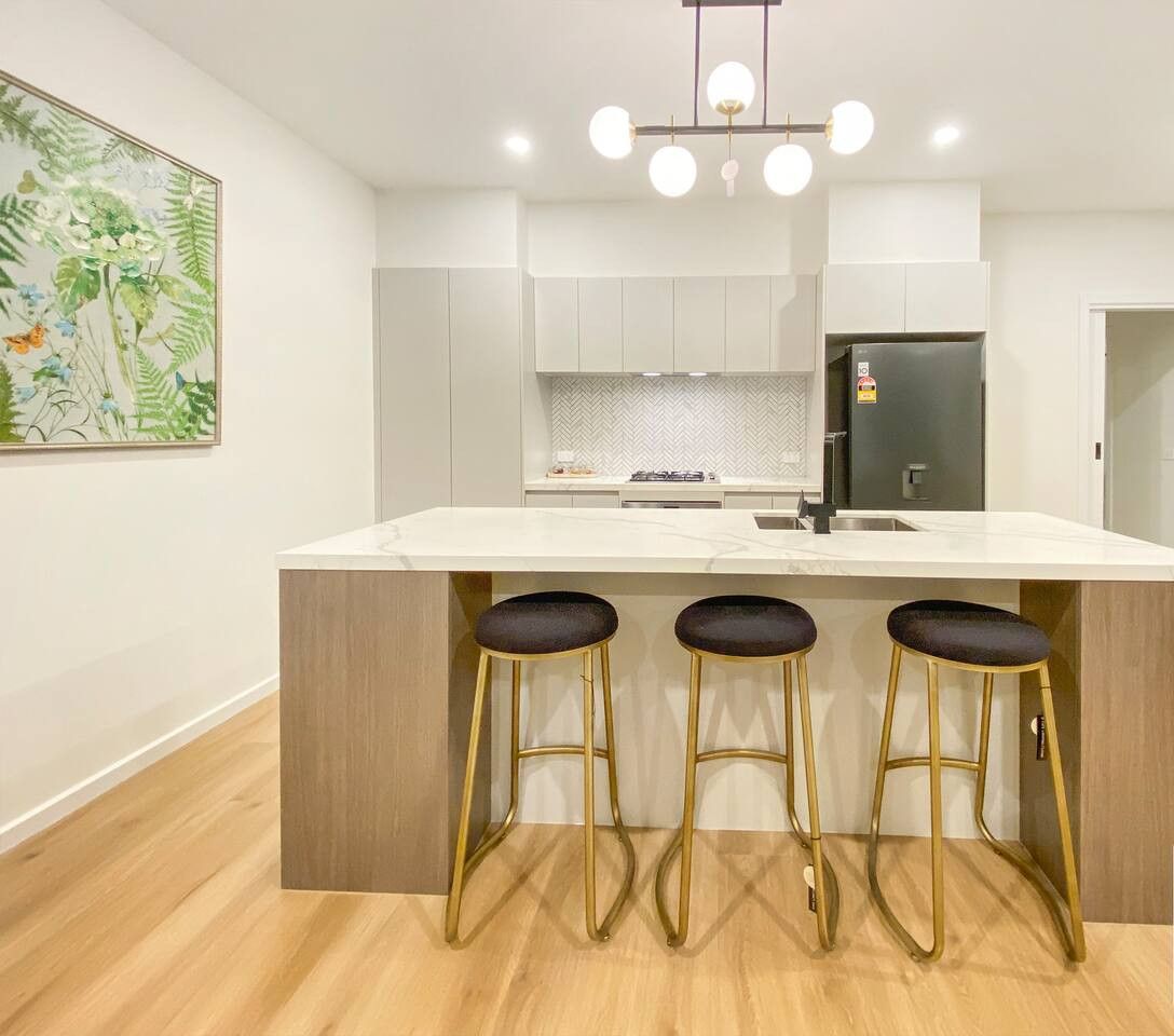 A modern kitchen with a marble countertop, three bar stools, a refrigerator, and a herringbone backsplash.