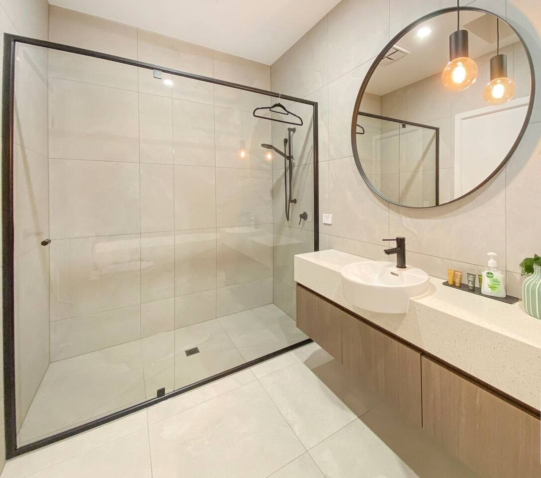 A modern bathroom with a glass-enclosed shower, a round mirror, and a vanity with a sink and toiletries.
