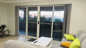 A living room with a grey sofa, a white coffee table, and a sliding glass door leading to an outdoor patio with a brick wall and chairs. - Modern, comfortable with stunning Inlet-sunset-view Home in the heart of Narooma (Narooma)