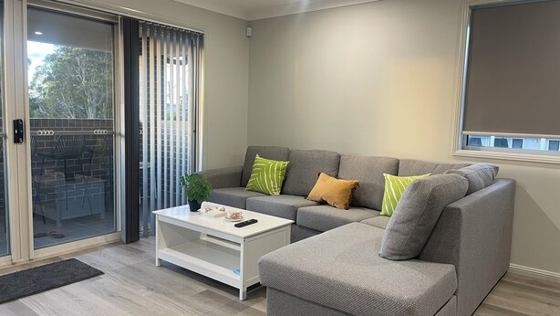 A modern living room with a grey sectional sofa, a white coffee table, and a sliding glass door leading to an outdoor patio. - Modern, comfortable with stunning Inlet-sunset-view Home in the heart of Narooma (Narooma)