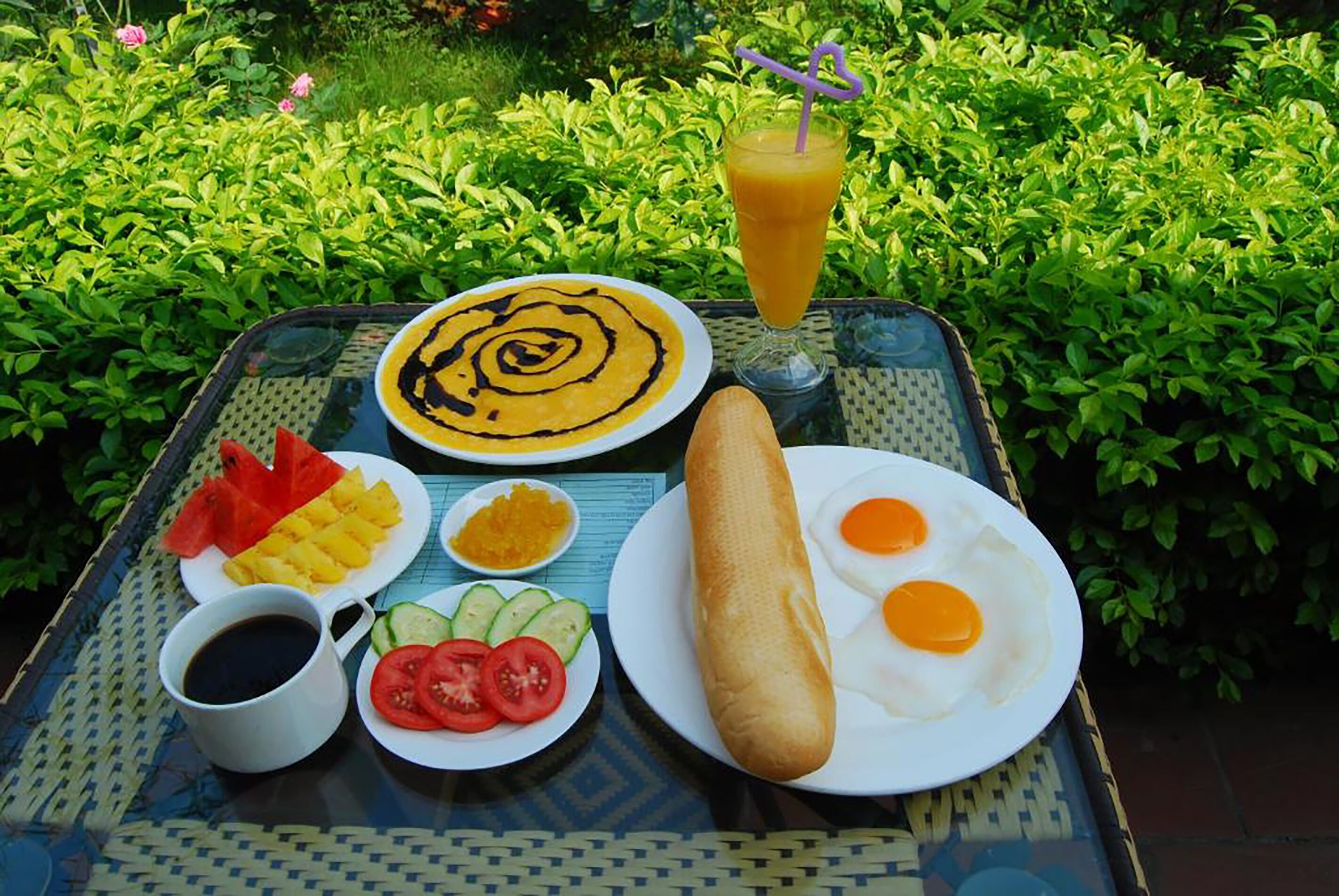 Uma refeição sobre a mesa com um copo de suco de laranja, um prato de ovos, uma baguete e acompanhamentos como tomates, pepinos e abacaxi.