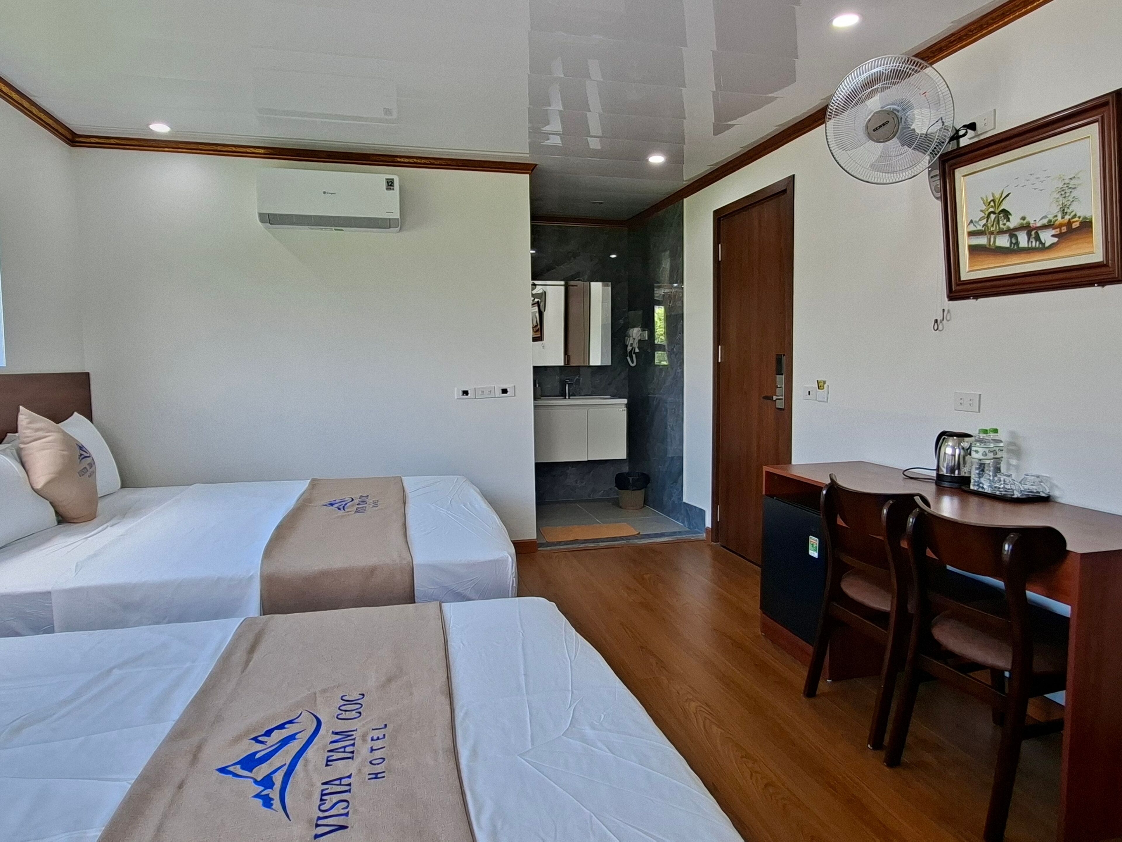 Camera d'albergo con due letti, una scrivania, una sedia, un ventilatore e un angolo cottura sullo sfondo.