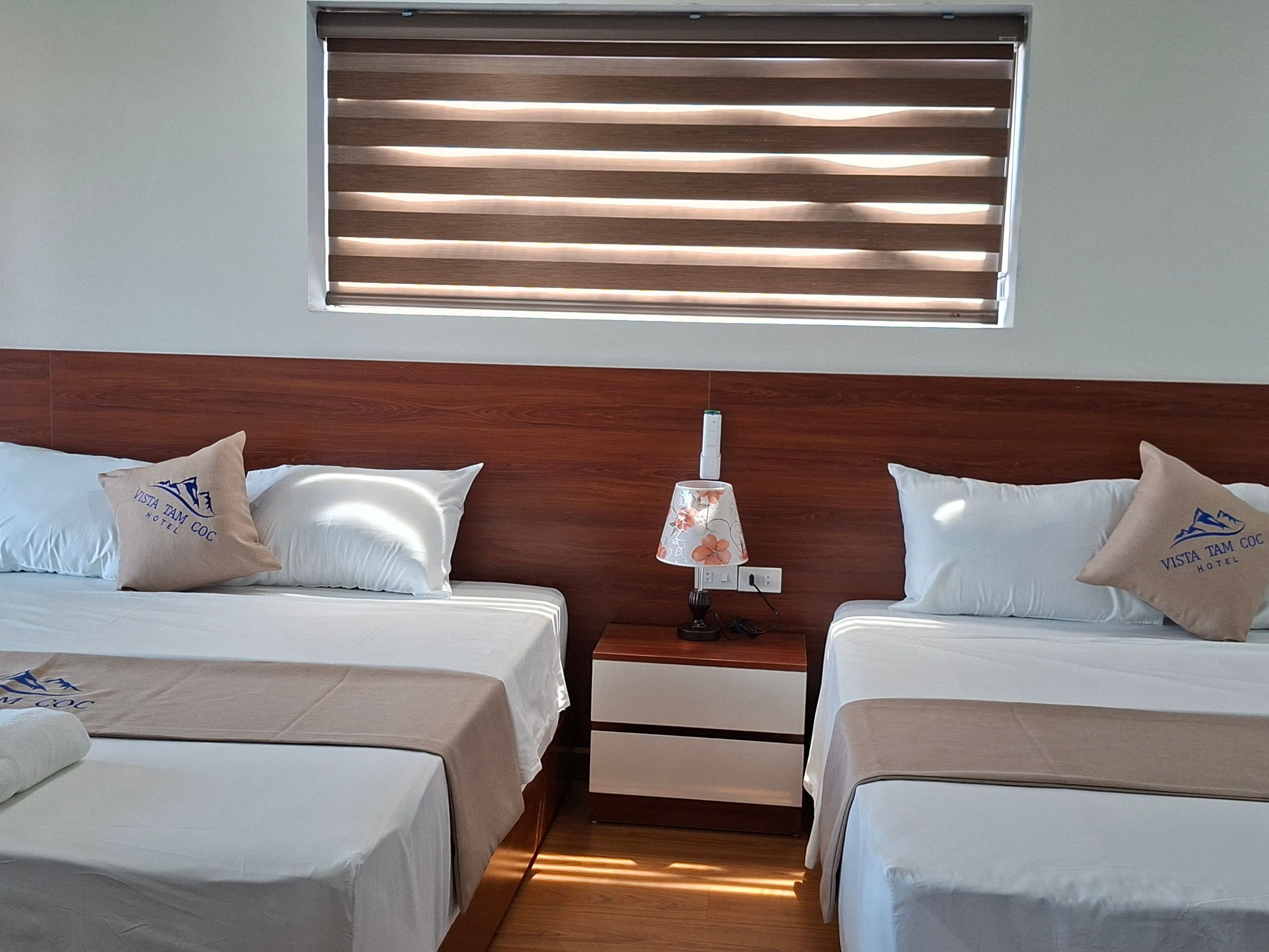 A hotel room with two beds, a wooden headboard, a nightstand with a lamp, and a window with blinds.