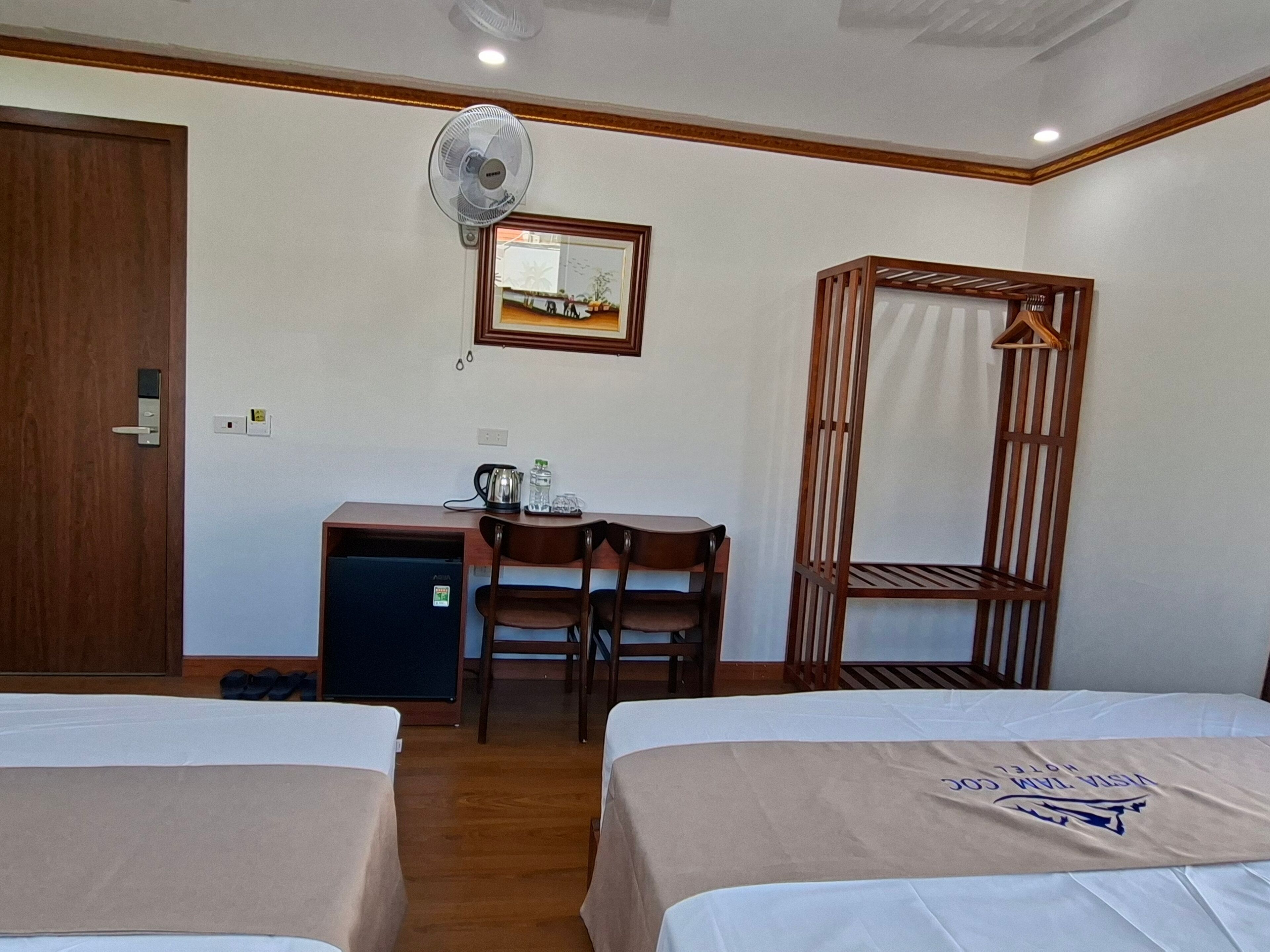 A hotel room with two beds, a desk with a chair, a small refrigerator, a wall-mounted fan, and a wooden wardrobe.