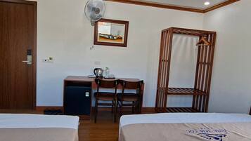 A hotel room with two beds, a desk with a chair, a small refrigerator, a wall-mounted fan, and a wooden wardrobe.