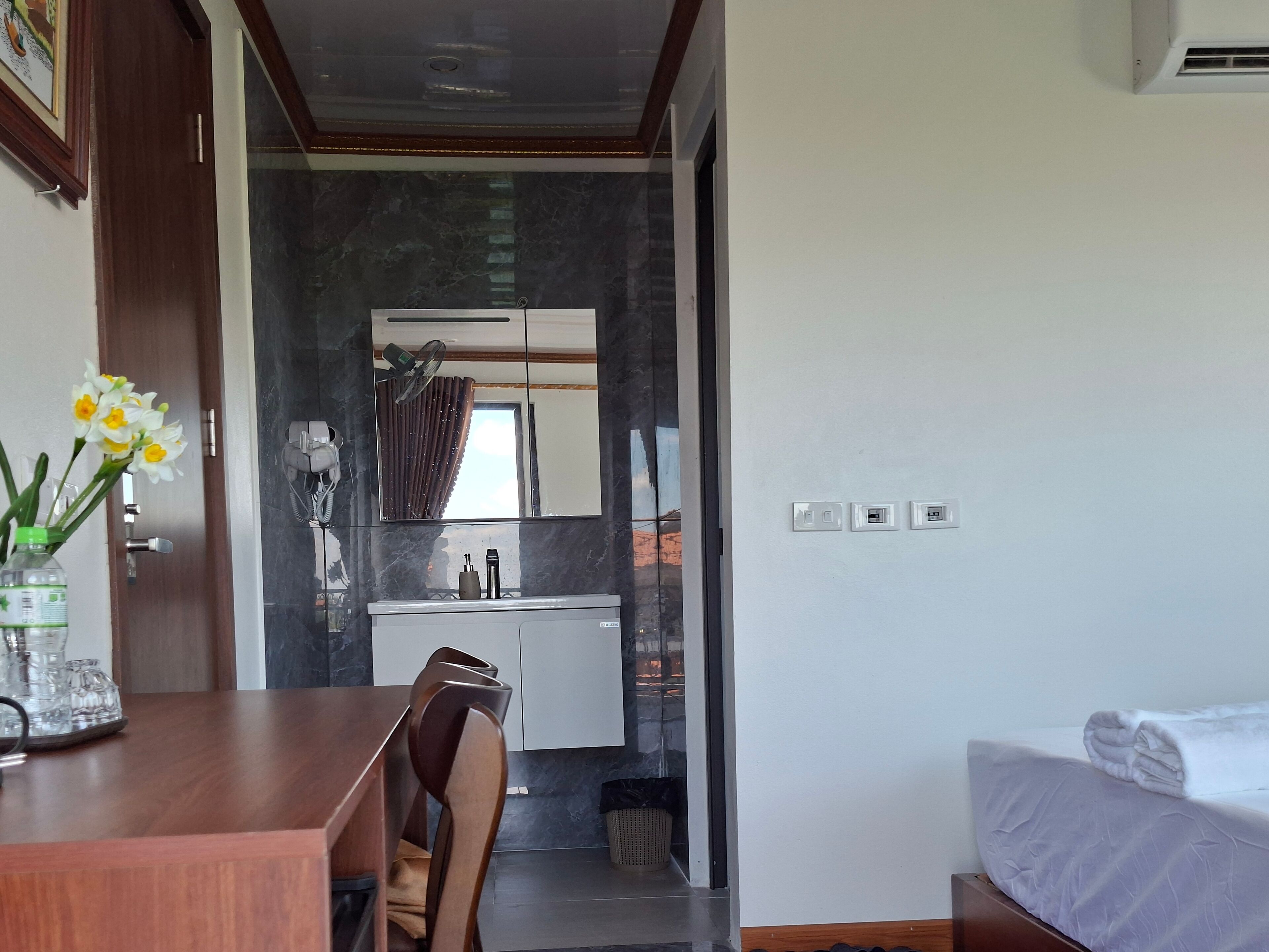 A modern hotel room with a wooden desk, a chair, a bed with a white comforter, and a bathroom visible through an open door.